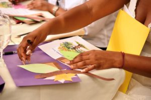 A person creating a craft project with colored paper leaves and ribbons, writing on a green leaf-shaped cutout, with other craft materials and a glass of water on the table.