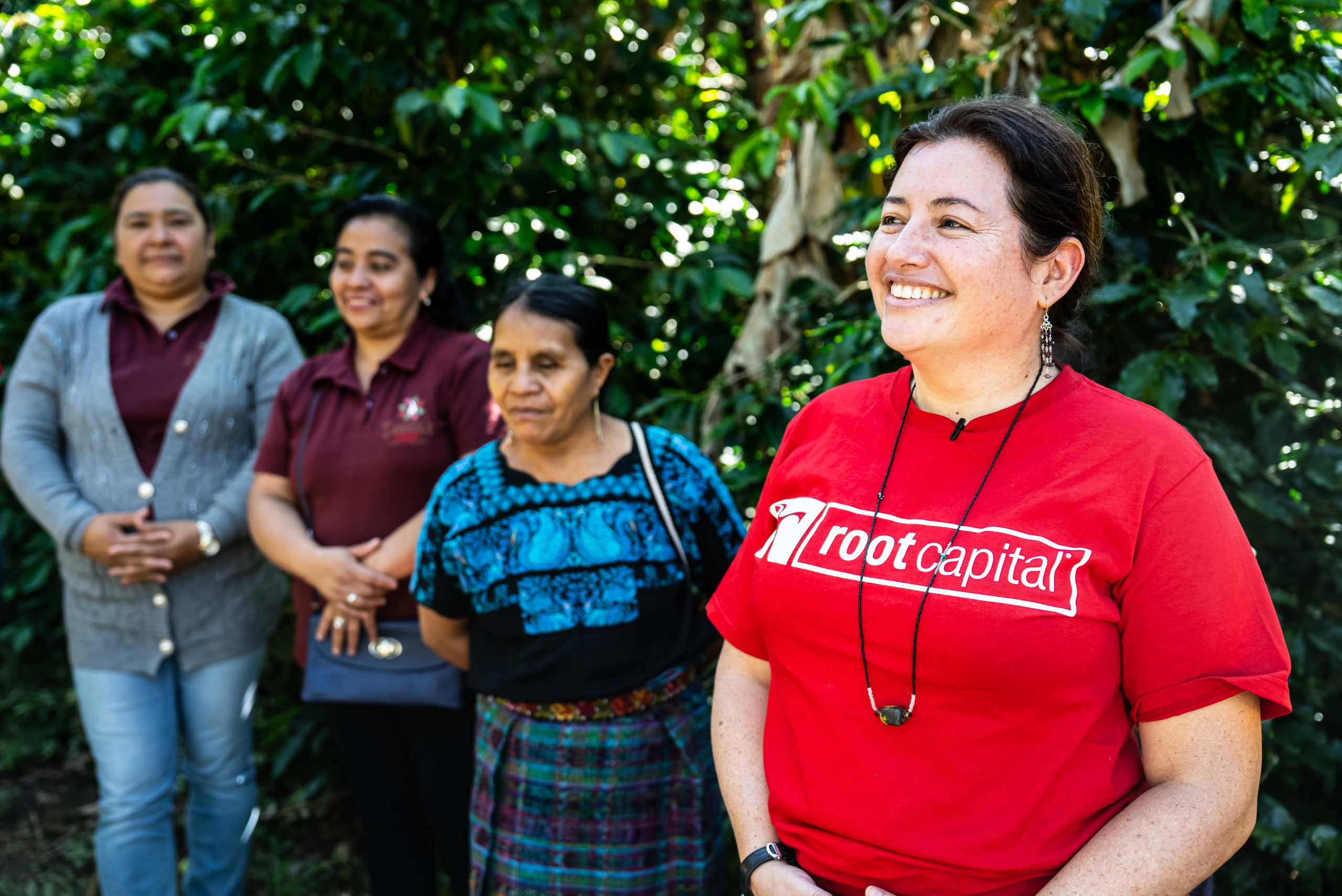 Four women stand outdoors in front of green foliage; the woman in front wears a red root capital shirt and smiles, while the three women behind her wear casual and traditional clothing.