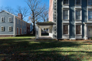 Two blue-grey historic wooden buildings connected by a covered walkway with white columns, surrounded by grass and bare trees on a sunny day. Shadows and sunlight create strong contrasts on the scene.