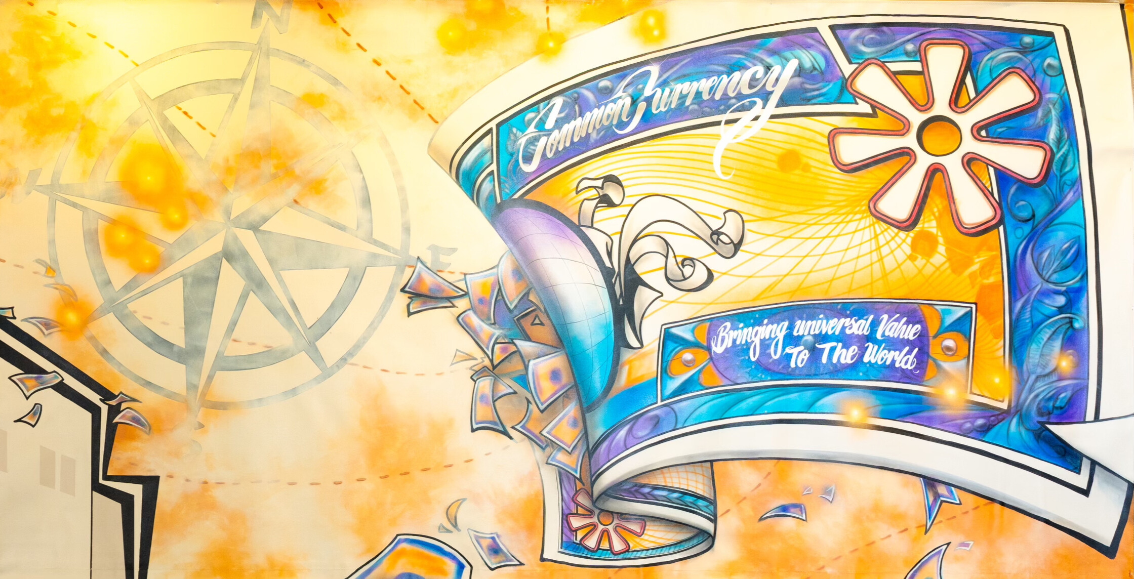 A vibrant mural features a large compass, abstract patterns, and a colorful banner that reads “Common Currency” and “Bringing universal Value To The World.” Yellow bursts and dynamic shapes add energy to the design.