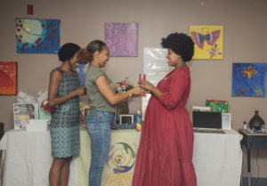 Three women stand together at a decorated table with colorful art on the wall. One woman in a red dress hands a candle to another woman, while the third woman watches and smiles. The setting looks warm and celebratory.