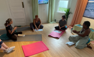Four women sit on yoga mats in a circle on a wooden floor, engaging in discussion with papers and books. The room has soft light, plants, and colorful curtains, creating a relaxed and inviting atmosphere.