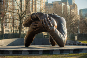 A large bronze sculpture of two arms hugging, without heads or bodies, set outdoors in a park with bare trees and city buildings in the background.