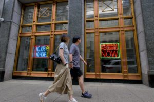 Two people walk past a building with large windows displaying neon signs that read, “NO JOB NO HOME” and “AFFORDABLE HOUSING NOW.” The street and building appear urban and modern.