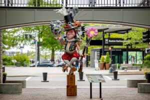 A whimsical statue of a grinning figure with a top hat, holding a flower and teapot, stands on tea crates under an archway near Boston Harbor Islands Welcome Center. Trees and street signs are visible in the background.