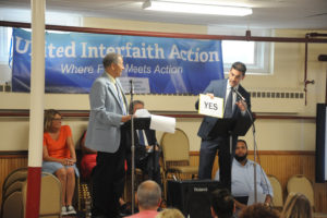 Two men stand at microphones in front of a United Interfaith Action banner; one holds a YES sign. Several people are seated and watching in the background in a community room setting.