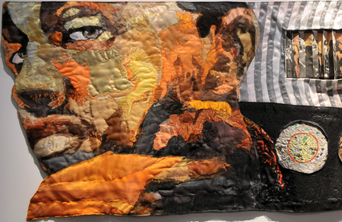 A colorful quilted textile artwork depicts a persons face in profile, made with various fabric patches in shades of orange, yellow, brown, and black. There are additional textured and patterned sections to the right.