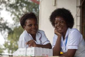 Two women in white uniforms smile while leaning on a counter with several medicine boxes in front of them. They appear to be healthcare workers, standing outdoors near a building.