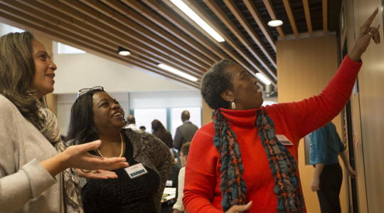 Three women stand indoors, smiling and engaged in conversation. One woman, wearing a red sweater and scarf, points at something on the wall while the others look on with interest. People and tables are visible in the background.