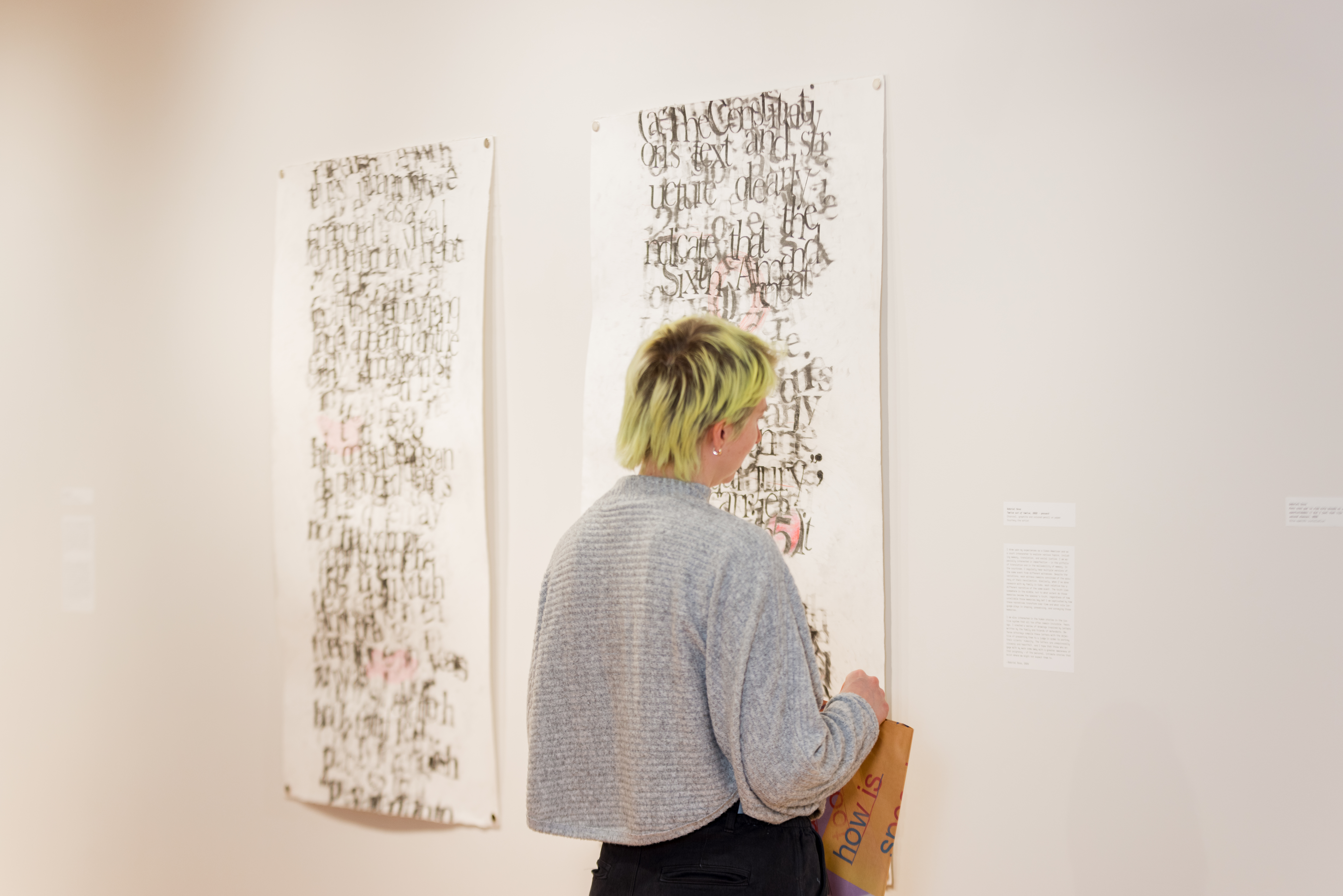 A person with short, light green hair views abstract, text-based artwork displayed on a gallery wall, holding a brown paper bag in one hand.