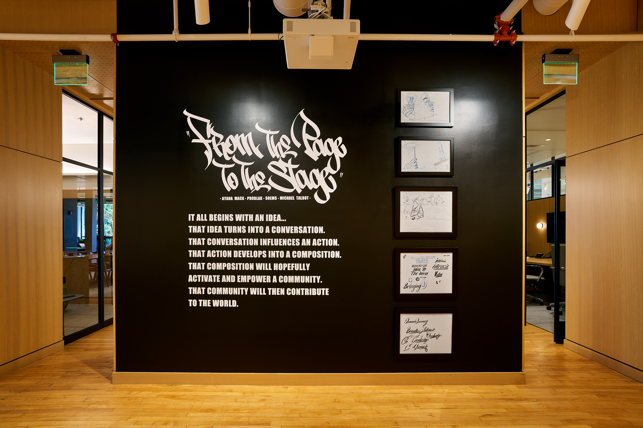 A modern office hallway features a black accent wall with white graffiti-style text reading “From the Page to the Stage,” followed by an inspirational quote and five framed handwritten notes or autographs. Light wood floors and open rooms flank the wall.