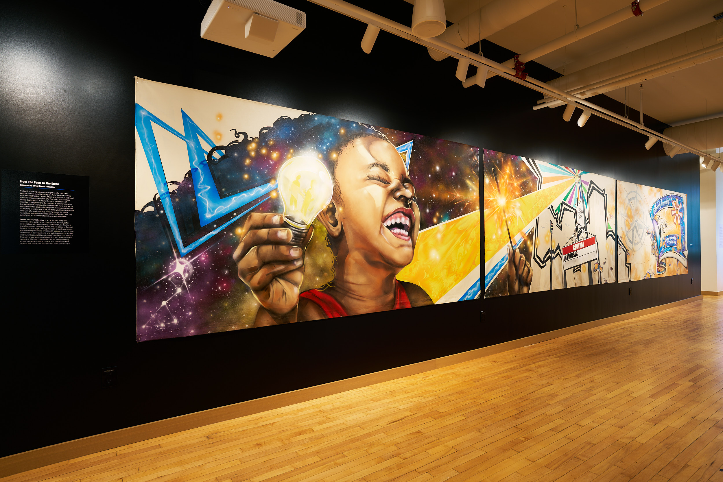 A vibrant mural on a black wall shows a smiling child holding a glowing lightbulb, surrounded by colorful, dynamic shapes and abstract patterns, with a wooden floor and overhead lighting in the gallery.