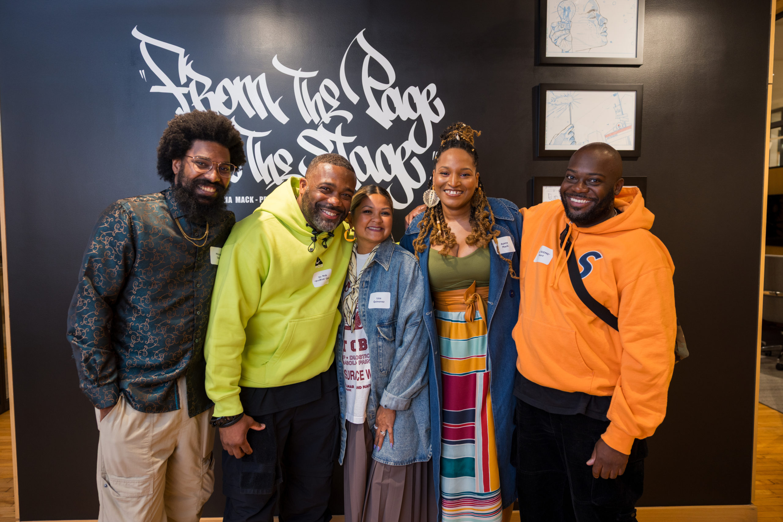 Five people stand smiling together indoors in front of a black wall with white graffiti text. They wear colorful, casual clothes and nametags. Framed artwork is displayed on the wall behind them.