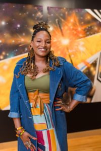 A smiling woman with locs wears a blue denim coat, green top, and colorful skirt, standing confidently in front of a vibrant, abstract artwork featuring yellow and orange tones.