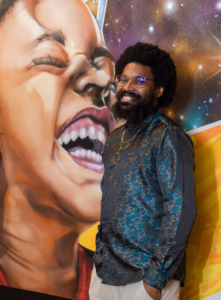 A man with glasses and a patterned shirt stands smiling in front of a colorful mural of a joyful person holding a glowing light bulb, set against a vibrant, cosmic background.