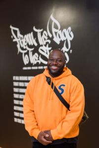 A man wearing an orange hoodie and a crossbody bag stands smiling in front of a black wall with white graffiti-style text that reads, From the Page to the Stage.