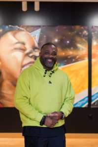 A man in a bright yellow hoodie stands smiling in a hallway with colorful, space-themed artwork depicting a child’s face on the wall behind him.