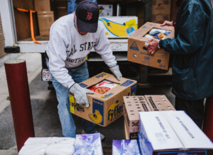 Two people unload boxes of bananas and other packaged goods from a truck. One person wears a cap, gloves, and a sweatshirt reading Real State North, while the other hands over a box.