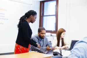 A woman stands showing something on a laptop to a seated man, while another woman works at a laptop in the background. All are in a bright office or classroom setting.