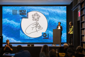 Two women stand towards the right of a stage behind a podium. A projected image fills a screen from the top to the bottom of the stage. The image is of a digital ocean-like background. In the center of this background is a white circle with a vector drawing of a hand holding a single flower. The petals of the flower are made of line drawings of eyes and ears.