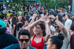 A large crowd of people dance outdoors at a festival or street event, smiling and enjoying the lively atmosphere. Trees, a fence, and a walk signal are visible in the background.