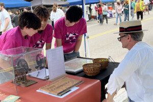 Three teens in matching pink shirts stand behind a table with artifacts and papers, talking to a man in a straw hat and glasses at an outdoor event with booths and people in the background.