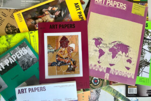 A variety of ART PAPERS magazine issues spread out, featuring colorful covers with artworks, maps, and text. The central issue displays an illustration of a person working on a craft or art piece.