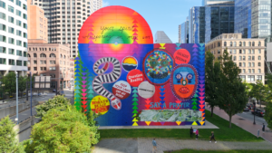 A colorful, vibrant mural on a city building features abstract shapes, faces, text like “Question Reality” and “Your Spirit is Showing in My Sky,” and vivid patterns, against a backdrop of tall modern and historic buildings.