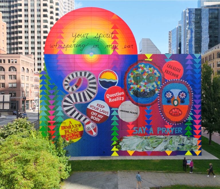A colorful mural on a city building features a large sun, flowers, a DNA strand, and bold text including “Your spirit whispering in my ear,” “Question Reality,” and “Keep Your Laws Off My Body.” Urban buildings surround the mural.