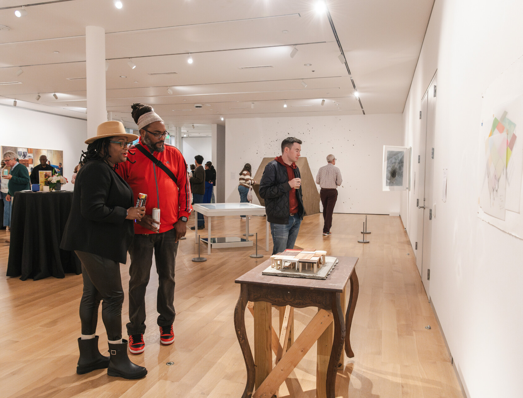 People view art exhibits in a modern gallery. Two individuals in the foreground examine a small architectural model on a table, while others observe artworks on the walls in the brightly lit space.