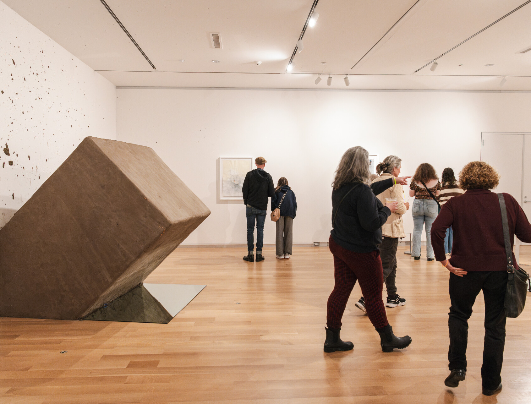 People view artwork in a modern gallery with wooden floors. A large, tilted geometric sculpture dominates the left. Several visitors observe framed art on the walls, while others converse in groups.