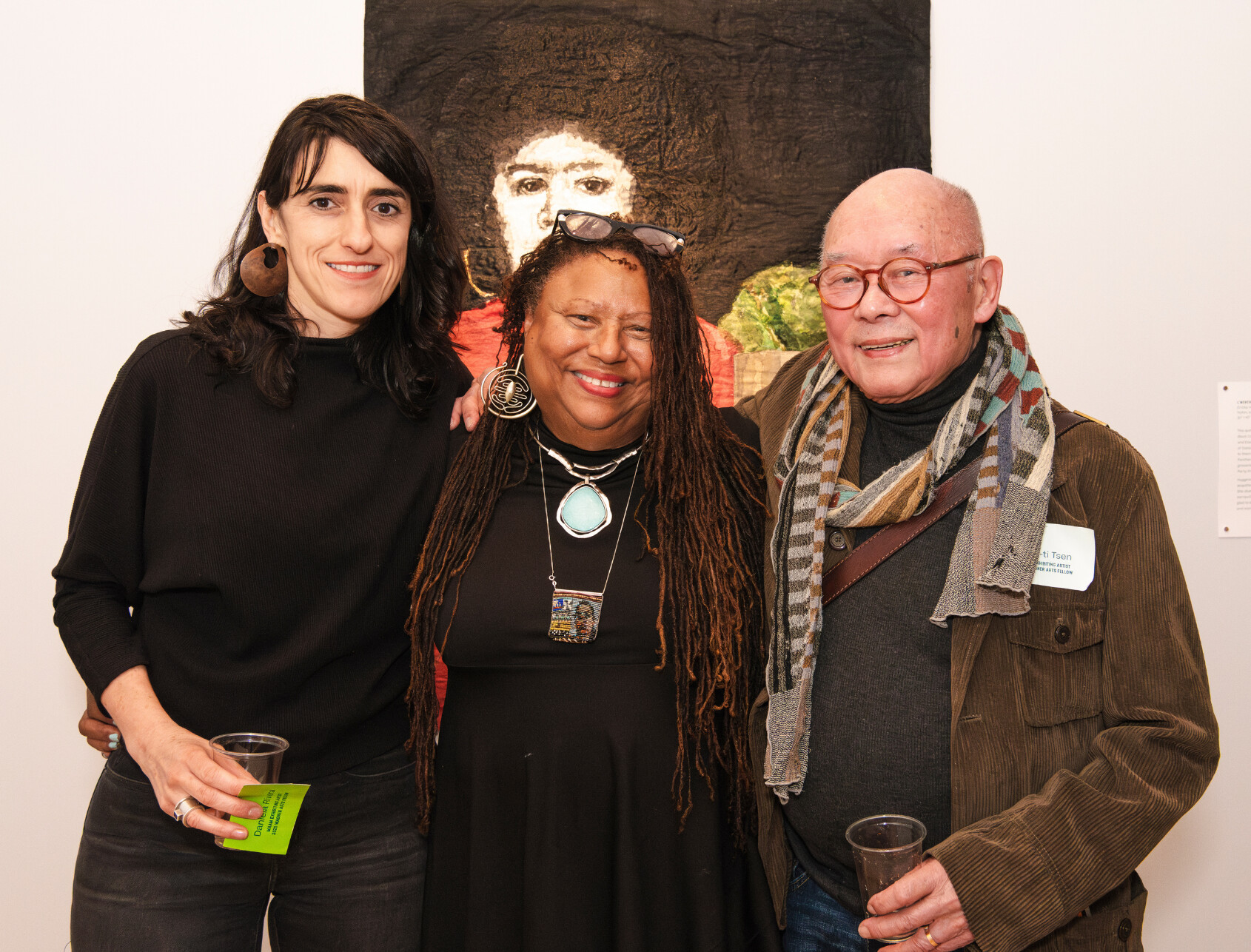Three adults stand together smiling at an art gallery, with a textured portrait hanging on the wall behind them. Two hold drinks, and one wears a scarf and glasses. All appear happy and relaxed.