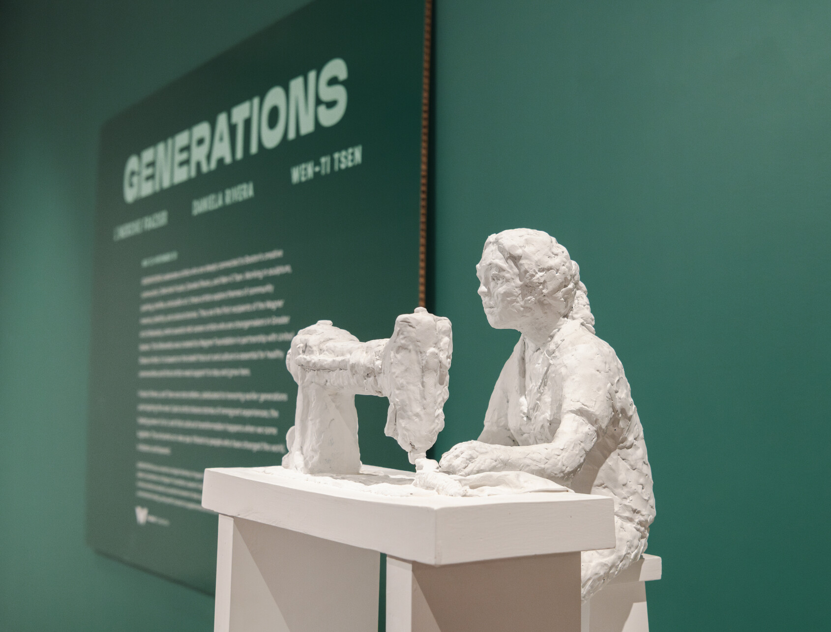 A white sculpture of a person using a sewing machine sits in front of a green wall with a sign titled GENERATIONS and descriptive text, featured in an art exhibit.