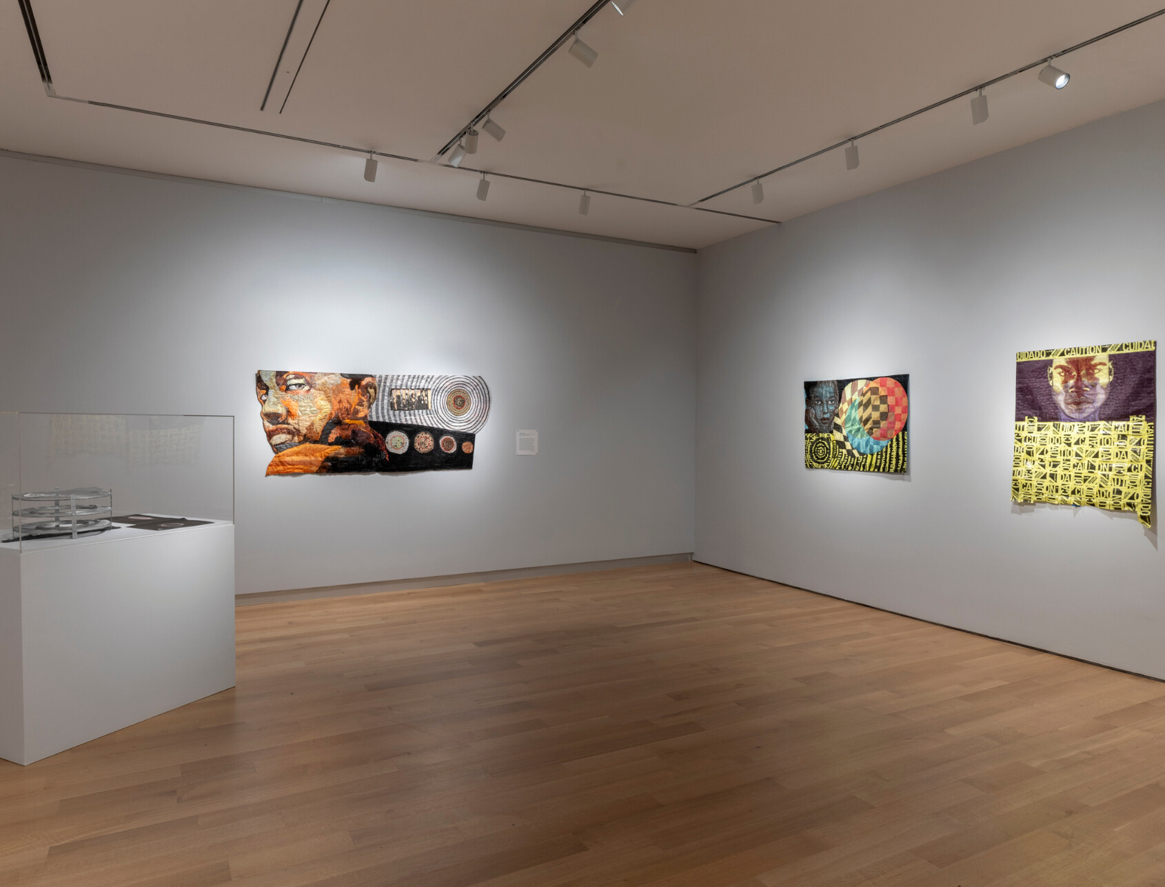 A modern art gallery with light wood flooring displays three colorful mixed-media artworks on a white wall and a white pedestal with glass objects on the left side. The room is brightly lit with ceiling spotlights.