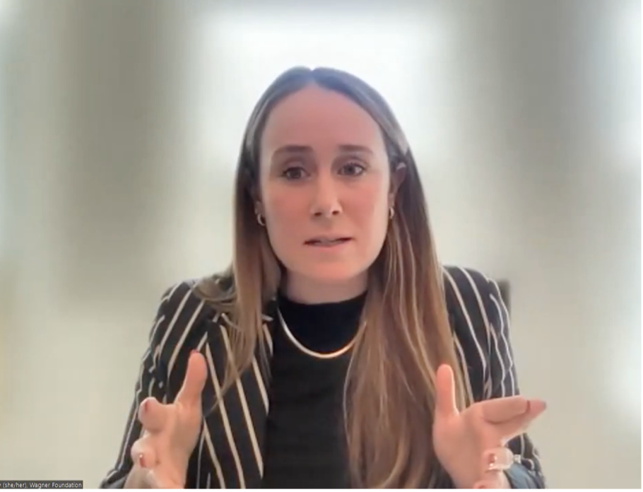 A woman with long brown hair, wearing a black and white striped blazer and gold necklace, gestures with her hands while speaking in a bright, softly-lit room.