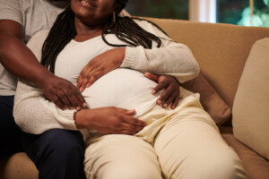 A pregnant woman reclines on a sofa while another person sits beside her, gently resting their hands on her baby bump. Both appear relaxed and affectionate.