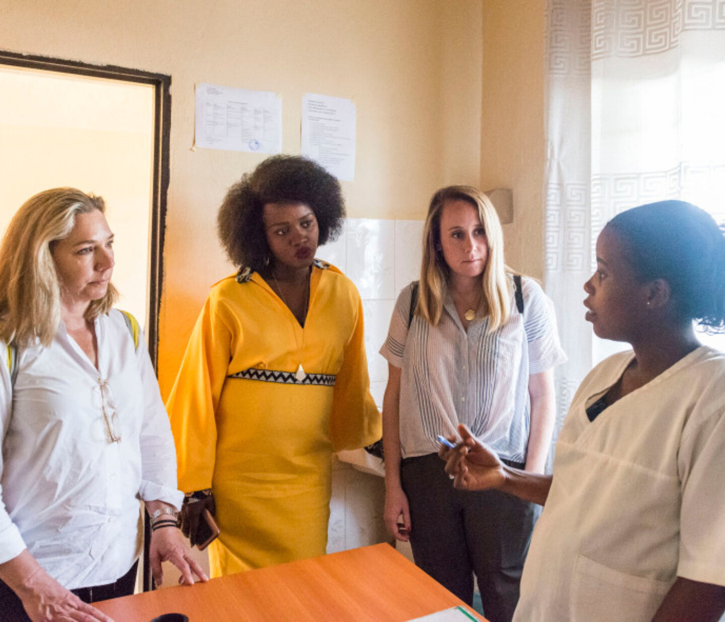 Four women stand together in a room, three listening intently to a fourth woman in a white uniform who appears to be explaining something. They are gathered around a wooden desk under natural light.
