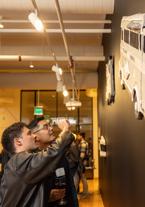 Two people examine and take a photo of 3D bus artwork mounted on a black wall in an art gallery, with other visitors visible in the background.