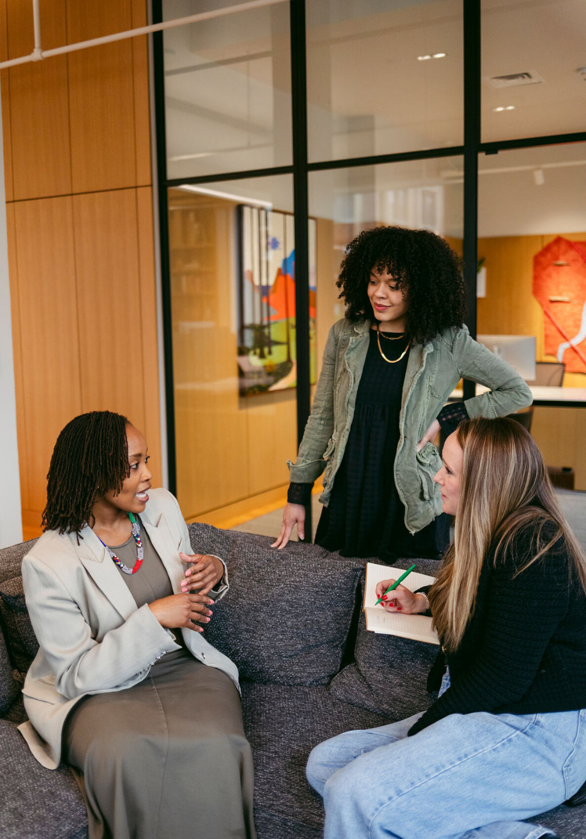Three women are having a discussion in a modern office. One is seated on a couch speaking, while the other two stand and listen attentively, one taking notes on a notepad.