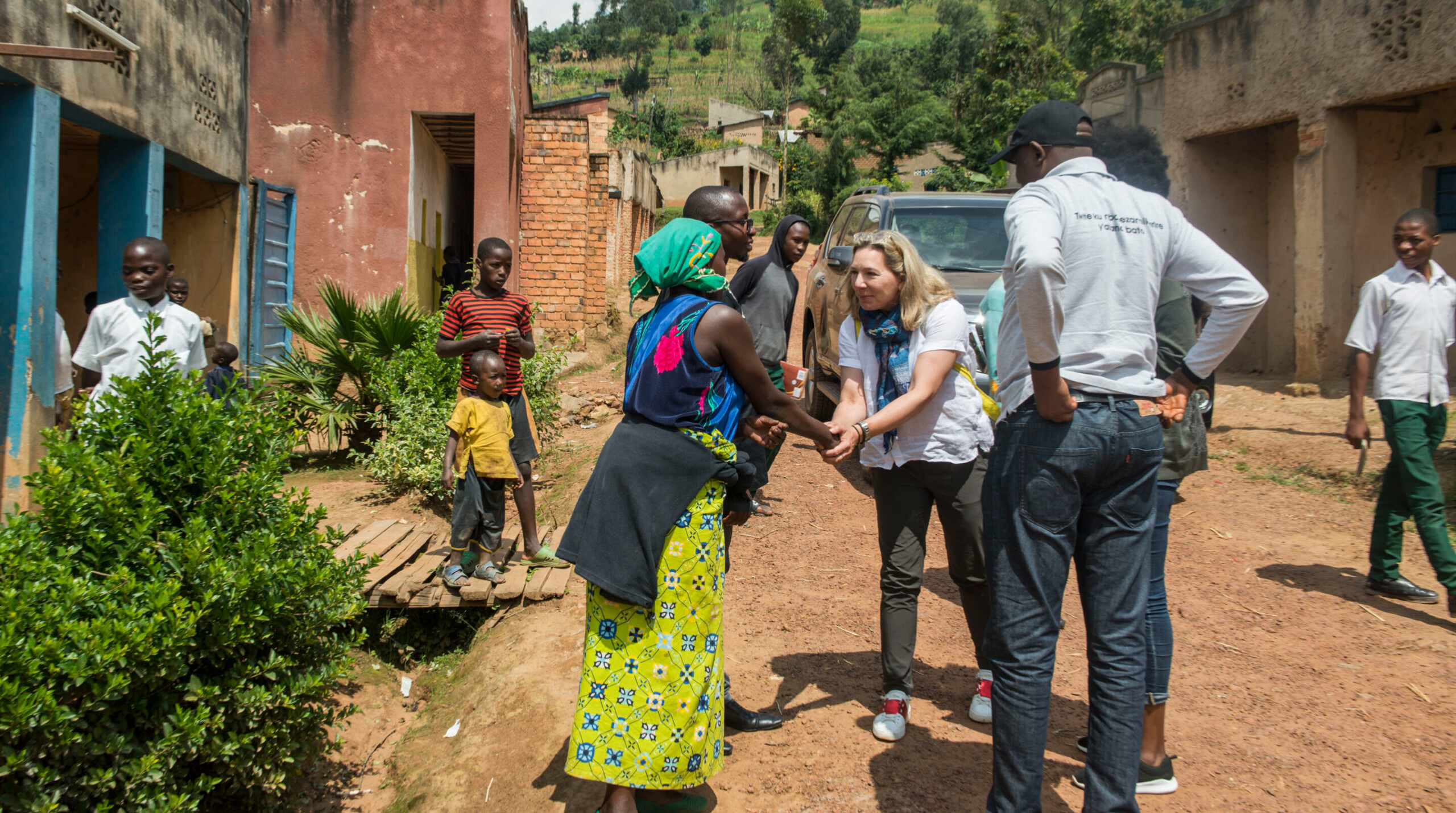 A woman in a green skirt and blue headscarf shakes hands with another woman in a white shirt, while several people stand and watch on a dirt road between buildings in a rural area.