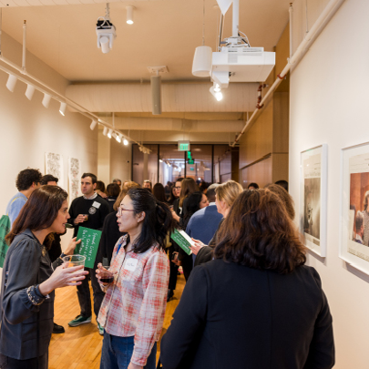 A group of people converse and hold drinks in a well-lit hallway during a social or networking event. Some people wear name tags, and the atmosphere appears lively and engaging.