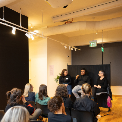 A group of people sit facing a panel of three speakers in a modern, well-lit room with wooden floors and black accents. One panelist gestures while speaking, and the audience listens attentively.