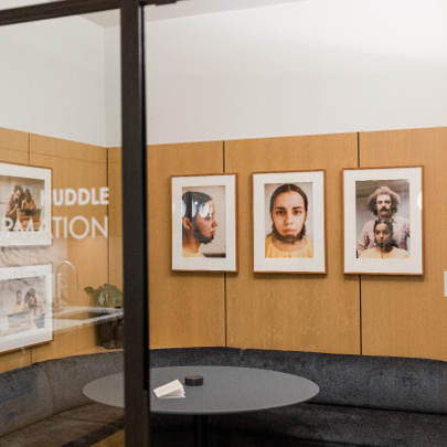 A small meeting room with light wood walls, a black round table, a curved gray sofa, and framed portraits of people hanging on the wall. The words HUDDLE and RATION are partially visible on the glass door.