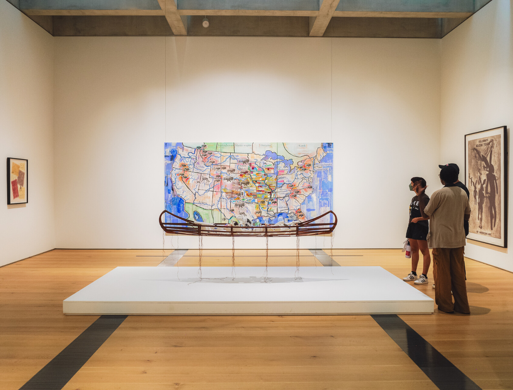 Two people view a museum art installation featuring a canoe-like sculpture in front of a large, colorful map on the wall; framed artworks hang on adjacent walls in the bright gallery space.