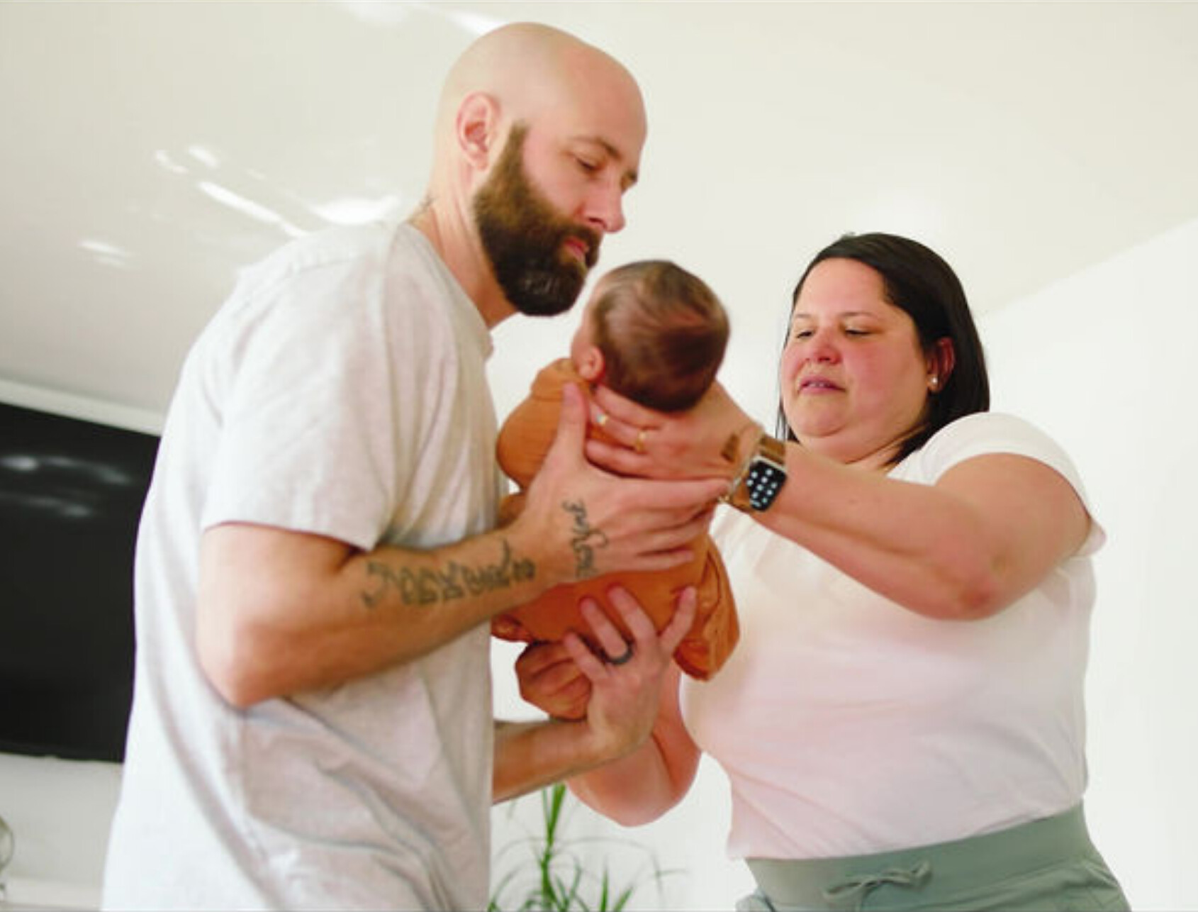 A bearded man and a woman gently pass a baby between them in a brightly lit room. The man wears a light t-shirt, and the woman wears a white top and smart watch. The baby is dressed in a brown onesie.