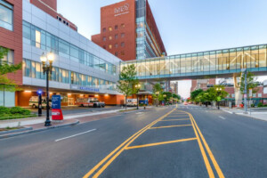 A wide city street is lined with modern hospital buildings. A glass skybridge connects two buildings above the road. Ambulances are parked outside under a sign reading EMERGENCY. The scene is quiet and well-lit.