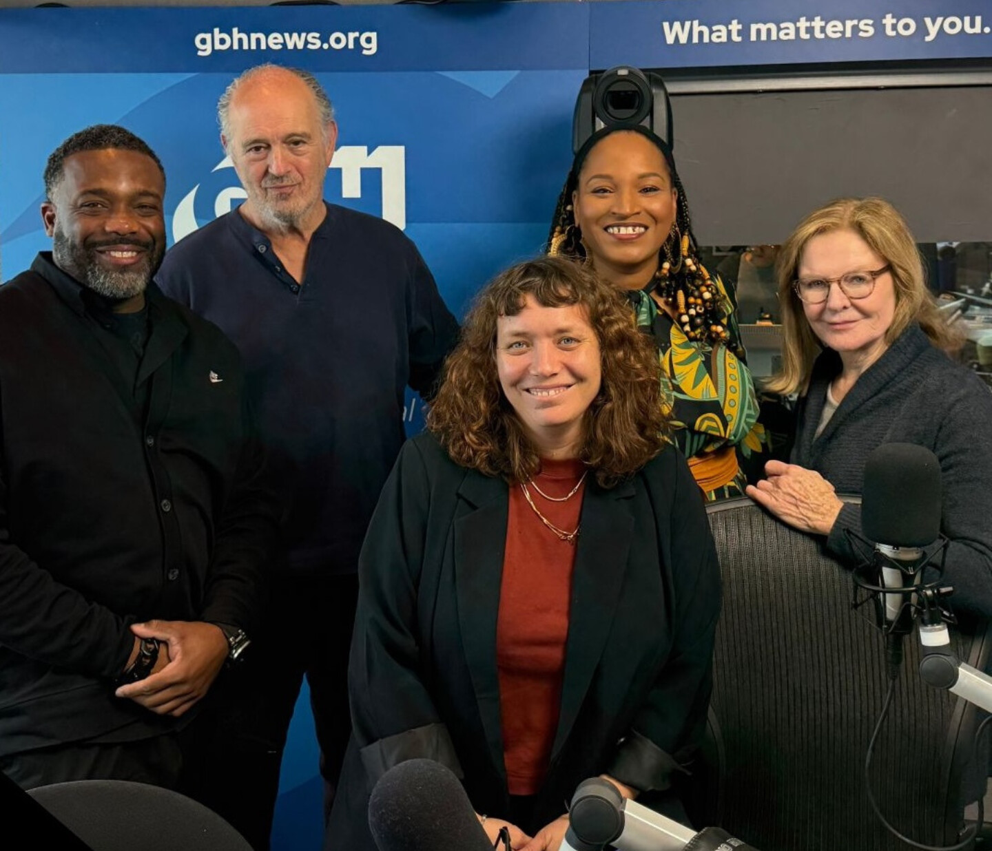 Five people stand and smile together in a radio studio, with microphones and a gbhnews.org sign in the background. Some are wearing dark clothing, and one person has a patterned outfit. The atmosphere appears friendly and professional.