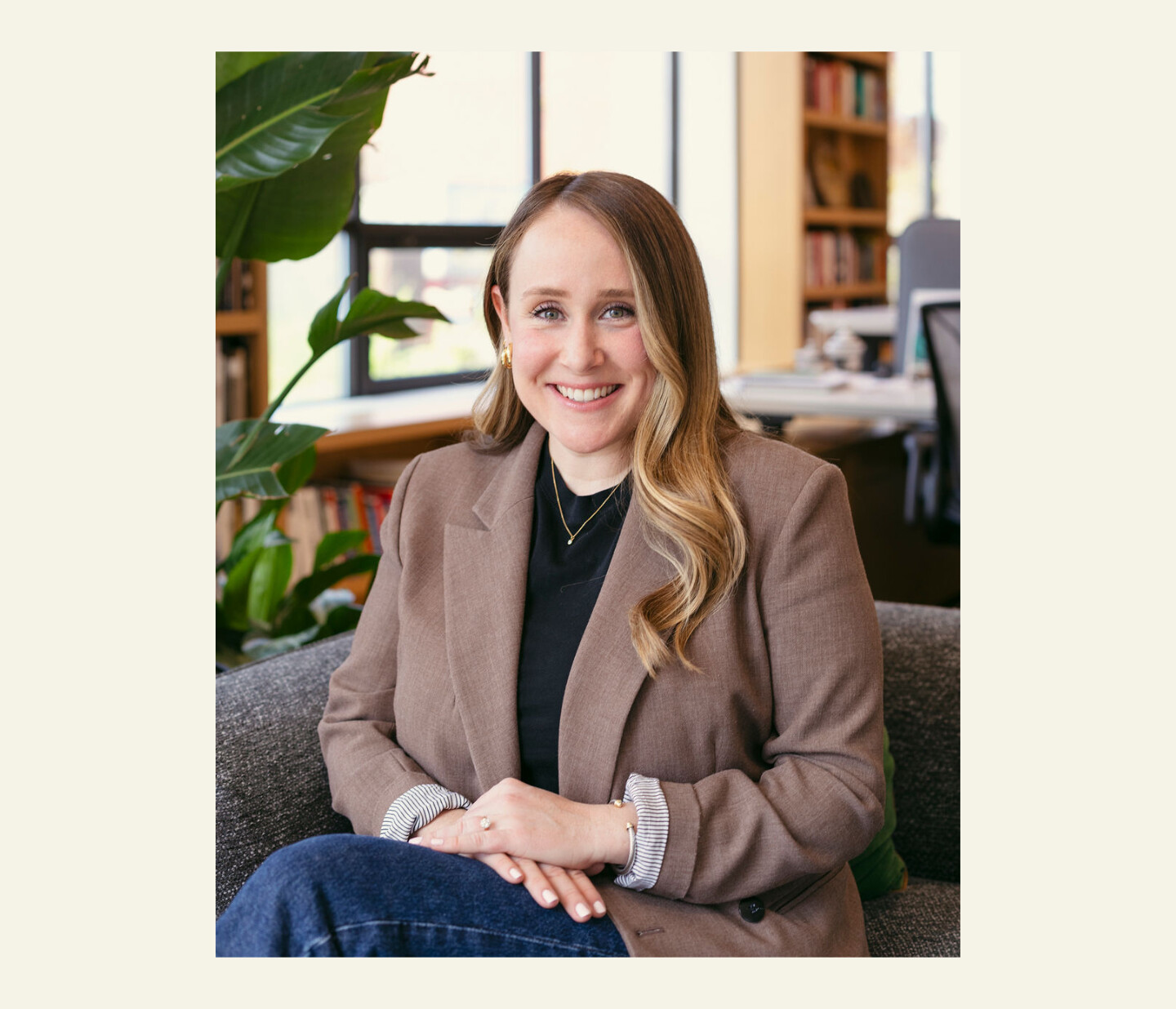 A woman with long, wavy blonde hair sits on a gray sofa, smiling. She wears a brown blazer over a black top. Bookshelves, a plant, and windows are visible in the background of a well-lit office.