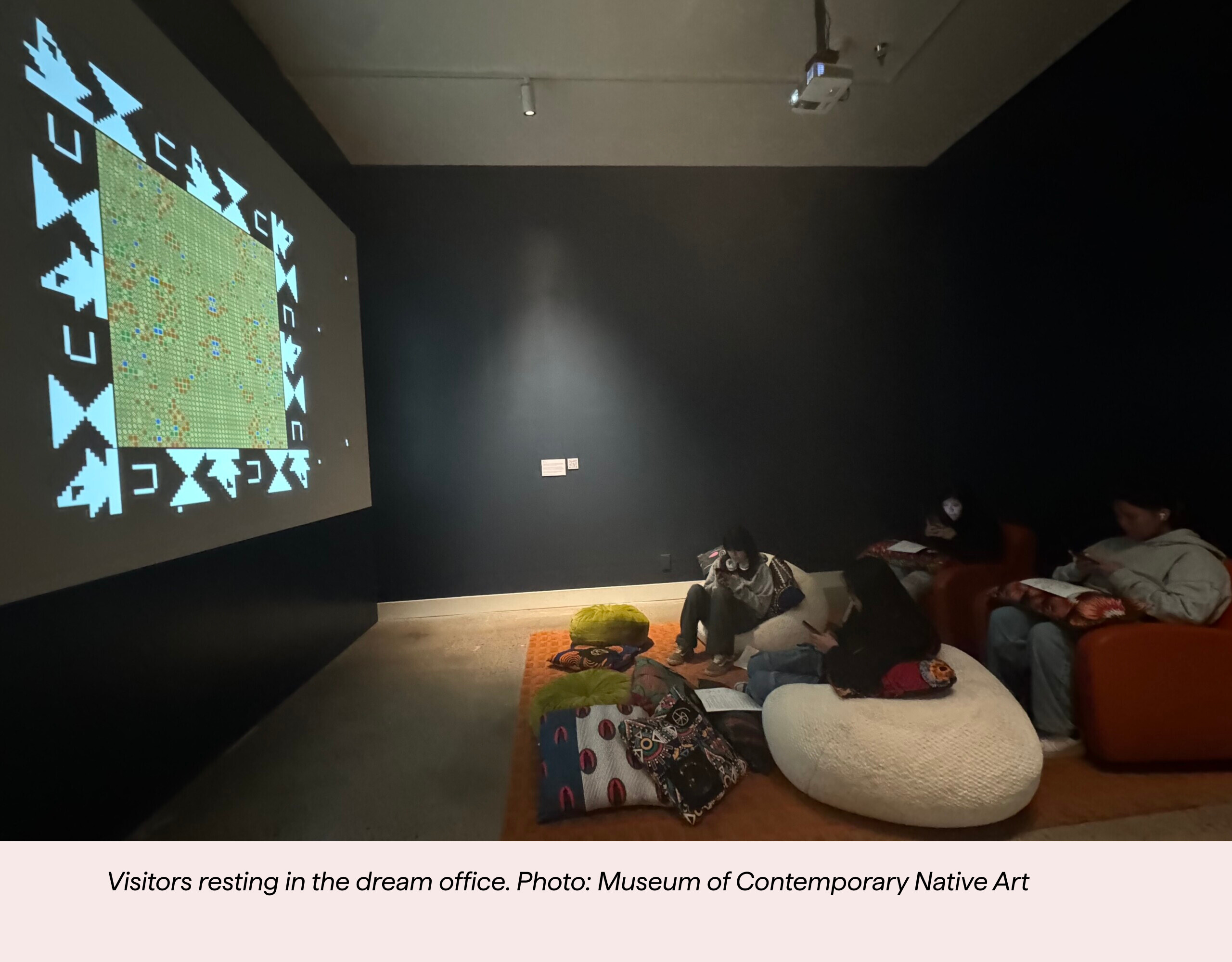 A dimly lit room with visitors relaxing on couches and cushions, watching a colorful, patterned projection on the wall. Blankets and pillows surround them, creating a cozy atmosphere.