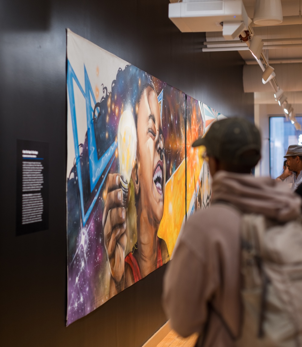 Two people view a vibrant mural in a gallery. The mural shows a smiling woman holding a glowing light bulb, surrounded by colorful and dynamic artistic elements on a black wall.